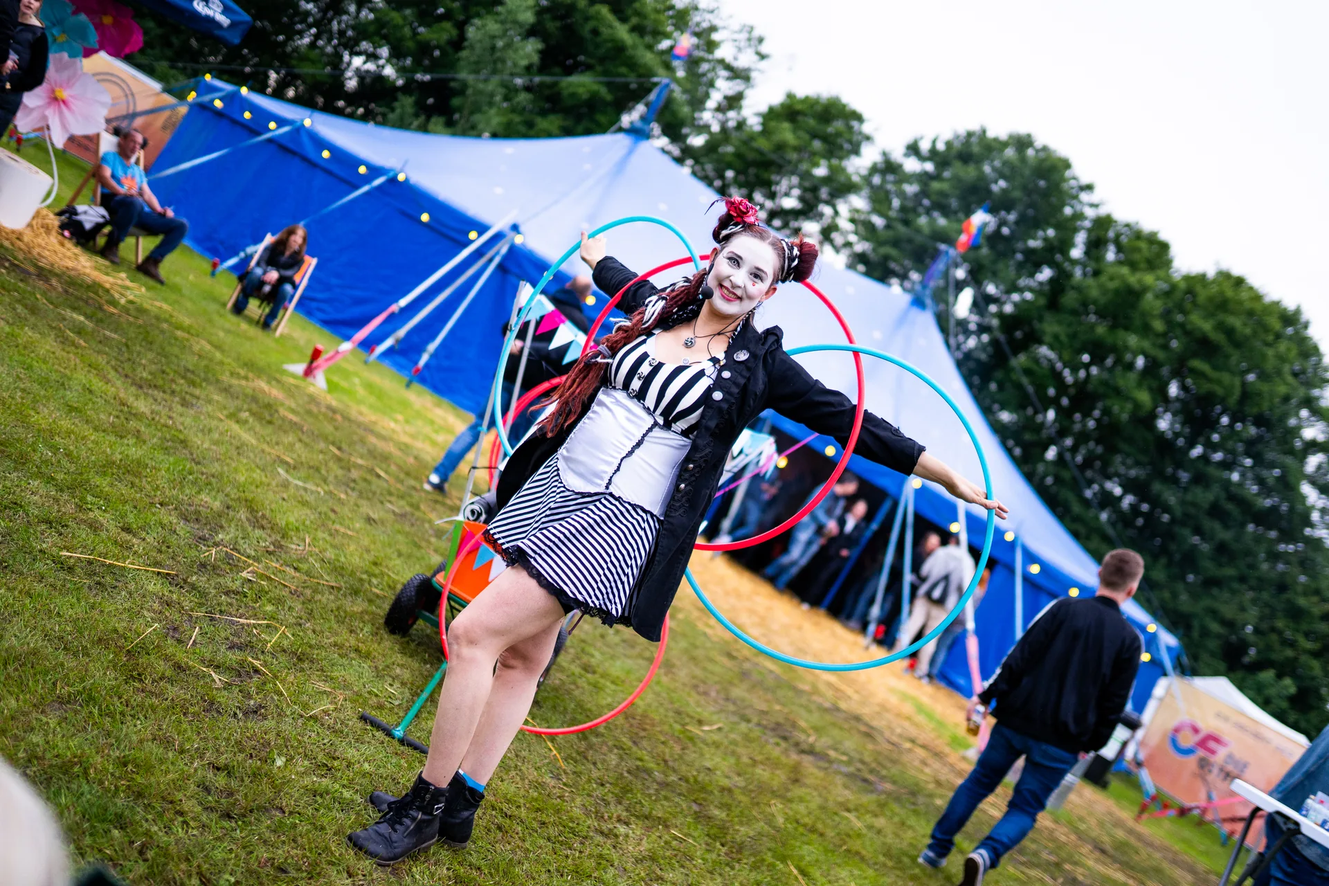 Interaktiver Circus-Clown Walk-Act mit Hula-Hoop-Reifen auf einem Festivalgelände vor einem blauen Zirkuszelt.