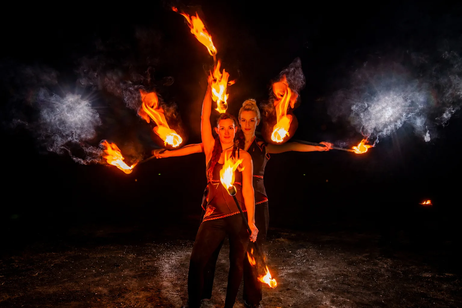 Zwei Feuerkünstlerinnen von Elements of Fire zeigen eine synchrone Partnerstaff-Performance bei Dunkelheit.